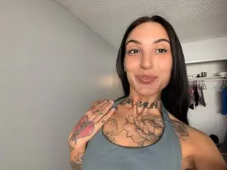 DylanAshley is a smiling woman with long dark hair and tattoos on her chest and shoulder, wearing a grey tank top.