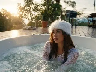 EIiMYJONES with long brown hair relaxing in a bubbling white hot tub outdoors during sunset.