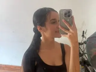 EMILYAlvernia poses close up, smiling while holding her phone, showing her long dark hair and black tank top against a plain white wall.