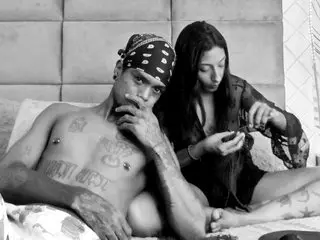 EbanoSpicyGirl poses shirtless with a hand to her mouth, showing off his tattoos and dark hair in a close-up shot.