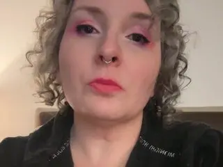 EdenArcadia has curly blonde hair and pink eyeshadow, wearing a black top while looking directly at the camera.