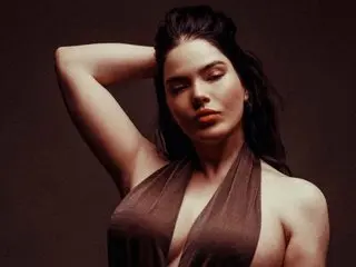 EdenEvansLive poses with her hands behind her head, wearing a low-cut brown top that shows her cleavage and shoulders against a dark background.