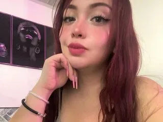 Eimy_Doll poses for a selfie with her hand under her chin, showing her long red hair and pink lipstick.