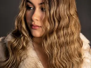 Ela_Curvy poses with her long, wavy blonde hair, looking off-camera with a soft expression against a dark background.