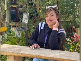 Elana_kepler smiles while posing outdoors, wearing a dark blue zip-up jacket with her dark hair down and hands resting near her face.