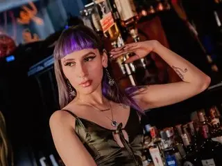ElectraRaptis poses with her hands behind her head, wearing a dark green satin top and showing her long purple hair with blunt bangs.