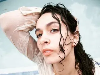 Electra_blue poses with her hand behind her head, showing her dark wavy hair and bare shoulders against a light blue background.