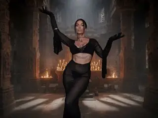 ElektraSheDemon poses in a black outfit, showing her cleavage and midriff, with her hands raised against a dark, moody background.