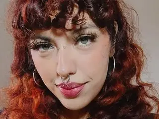 EliFlores looks at the camera with her curly red hair, nose ring, and hoop earrings, showing her face and light makeup.