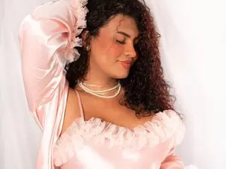 EliizabethSantos poses in a pink satin top with ruffled trim, showing her dark curly hair and bare shoulders against a white background.