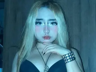 ElizabethCrowleyy with long blonde hair, wearing a black low-cut top, posing against a dark background.