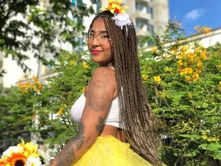 Elizabetth_Romann smiles while posing outdoors in a white top and yellow skirt with flowers in her long braids.