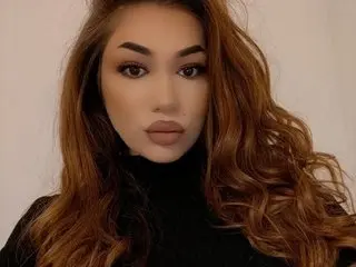 EllaBelleX poses with long wavy brown hair and a black turtleneck against a plain white background.