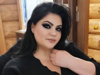 EllaXJane poses with her long black hair and dark eye makeup, wearing a black top while resting her hand near her neck.