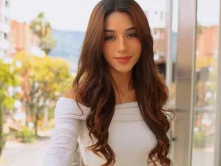 Ellen_Harris poses with long wavy brown hair, wearing a white off-the-shoulder top against a soft outdoor background.