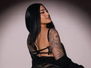 Tattooed woman in a black sheer outfit posing elegantly against a soft studio background.
