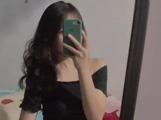 ElysiaBae poses for a mirror selfie, showing her long dark wavy hair and black off-the-shoulder top in a dimly lit room.