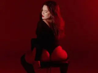 EmaBaaker poses with long dark hair, wearing a black long-sleeved top and red bottoms against a dark red background.