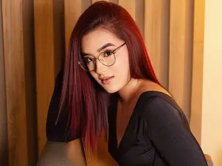 EmaLane poses with long red hair and glasses, wearing a black top against a warm, wooden paneled background.