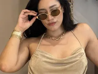 EmaLenox wears gold sunglasses and a gold necklace while posing in a beige halter top.