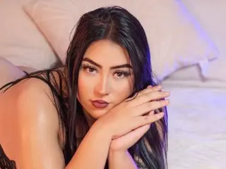 Emberrvelvet poses with her hands near her face, showing her long dark hair and bare shoulders against a soft white pillow.