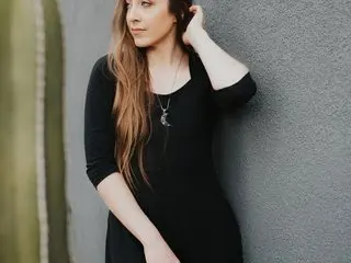 EmeraldOstara poses with her long brown hair, wearing a black long-sleeved top against a plain wall background.