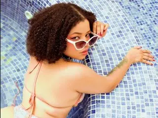 Emiilycollins18 with curly dark hair poses in a pink bikini against a blue tiled pool background.