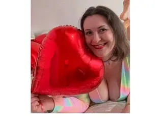 Smiling woman in a colorful pastel top holding a large red heart balloon in a cozy indoor setting.
