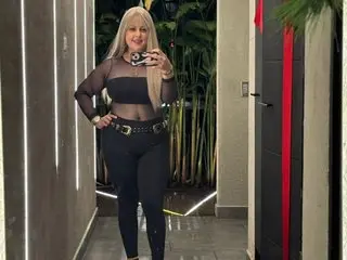 EmillyRosse poses in a black mesh top and black leggings, showing her blonde hair and figure against a dark indoor background.