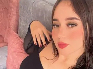 EmilyBrown199 with long dark hair and red lipstick poses on a grey couch wearing a black top.