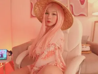 EmilyChachati with long pink hair wearing a straw hat and a pink floral outfit while sitting in a white chair.