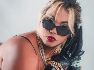 EmilyDuran99 poses with blonde hair and black sunglasses, wearing a black strap and heavy gold chains against a light background.