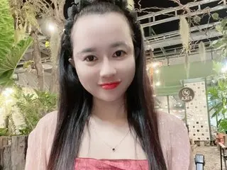 EmilyShow_96 smiles at the camera with long black hair and red lipstick, wearing a pink top in an outdoor garden setting.