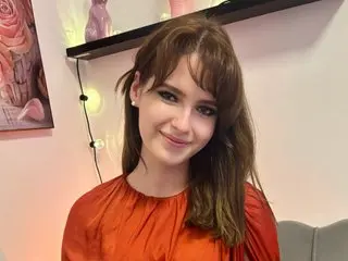 Emily_Paris smiling with long brown hair and bangs, wearing a red silk blouse against a plain white wall.