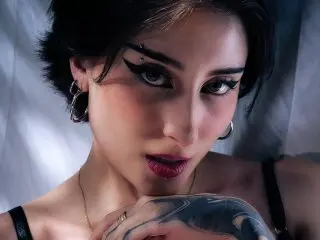 EmmaCollins67 poses with a direct gaze, showing her dark hair, heavy eyeliner, and hoop earrings against a soft, light background.