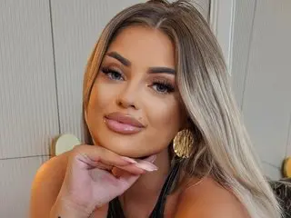 EmmaMavis poses with her hand under her chin, showing off her long blonde hair and makeup in a close-up shot.