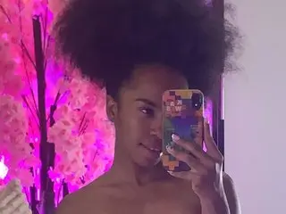 Emma_Lovve poses shirtless with her dark, voluminous hair, looking directly at the camera in a setting with bright pink lighting.