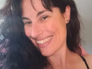 Smiling woman with long dark wavy hair in a casual black tank top indoors.