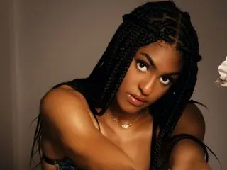 EmoriSplash poses with her long black braids, showing her bare shoulders and chest against a plain dark background.