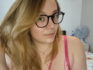Emski_xox poses close-up wearing black glasses and a pink strap, showing her blonde hair and face in a bright room.