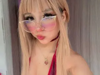 EmyGom3zz poses with long blonde hair and pink-tinted glasses, wearing a red top that shows her cleavage.