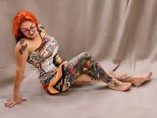 Red-haired woman in a patterned floral jumpsuit sitting on a grey studio floor with a relaxed mood.