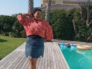 EnigmaticGoddess stands outdoors by a swimming pool wearing a red patterned button-down shirt and a denim skirt, running her hands through her short dark hair.