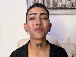 Eri_hott looks directly at the camera with a slight smile, showing her short dark hair and a neck tattoo, wearing a black top against a plain wall.