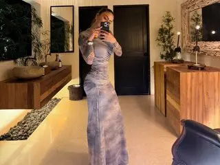 EricaCopolaa poses in a long, tight grey dress, holding her phone to take a mirror selfie in a room with a wooden console and dark door.
