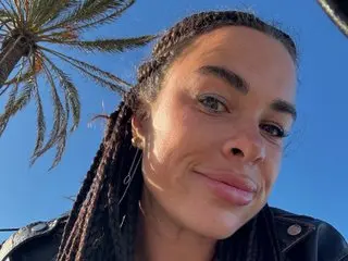 ErikaCutess smiles outdoors under a blue sky with palm trees, wearing a black leather jacket and showing her long dark braids.