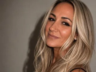 Esme_xxx smiles at the camera with long blonde hair against a grey wall.