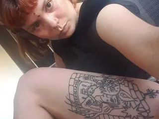 Redheaded woman in a black tank top showing a large black ink tattoo on her thigh in a casual indoor setting.