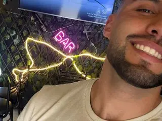 Ethan_Vance smiles while posing close-up, showing his face, dark hair, and beard in a dimly lit setting.