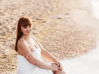 EvaSaint has long red hair and wears a white strap dress while sitting on a sandy beach.